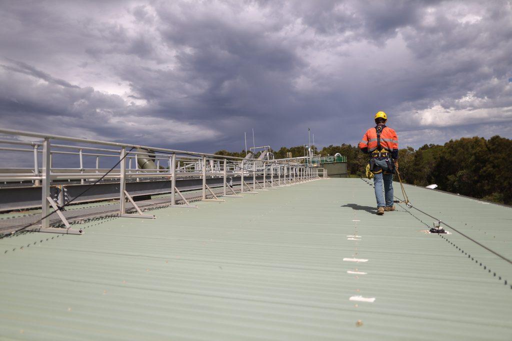 Construction safety inspector wearing fall safety harness walking on the roof by clipping safety rope on fall arrest, fall restraint horizontal safety line while inspecting fixed roof anchor points