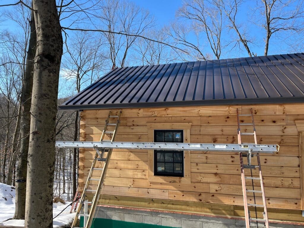 newly built cabin in snow with standing seam metal roof with ladders leaning against wooden side